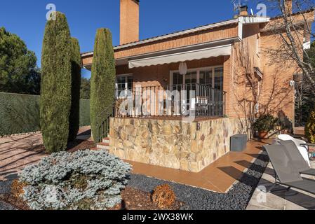 Chalet staccato con pavimento in ghiaia, alcuni cipressi all'interno, portico con ringhiera metallica, piante e siepi Foto Stock