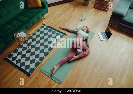 Una donna in abbigliamento sportivo si stende su un tappetino yoga sul pavimento in legno duro di un soggiorno e si allunga Foto Stock
