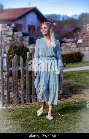 Una giovane donna dai capelli biondi e un sorriso radioso passeggia per il parco in una giornata di primavera assolata. Indossa un vestito alla moda che si mette in mostra Foto Stock