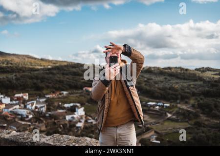 Giovane e felice fotografo che viaggia in visita turistica in una piccola città, scattando una foto con il suo cellulare della vista panoramica della città e del monte Foto Stock