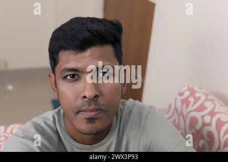 Un uomo indiano premuroso siede su un divano, guardando direttamente lo spettatore con un'espressione seria Foto Stock