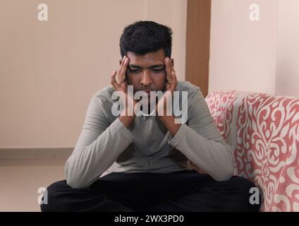 Un uomo indiano siede su un divano nel suo salotto, massaggiando i templi nel dolore causato da un mal di testa. Foto Stock