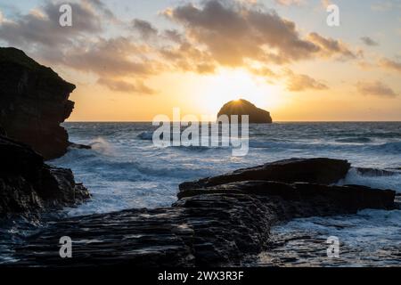 The sun setting behind Gull Rock viewed from Trebarwith Strand Beach, North Cornwall UK. Evening scene with waves, surf and jagged cliffs. Foto Stock