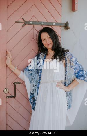 Una donna sorridente con un abito bianco e uno scialle blu si appoggia su una porta in legno rosa, trasudando eleganza e calore casual. Perfetto per lo stile di vita Foto Stock