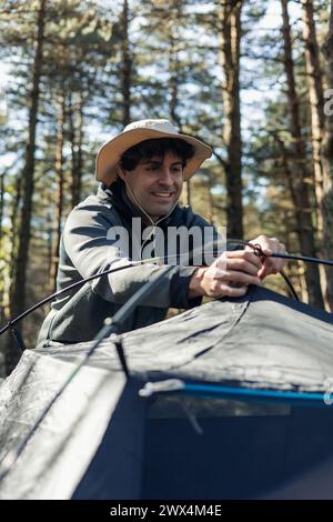 Primo piano di un giovane sorridente che indossa un cappello, sistemando la sua tenda in mezzo alla bellezza serena del campeggio durante l'ora d'oro del tramonto su una somma Foto Stock