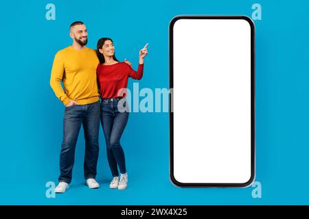 Coppia sorridente in piedi accanto al telefono grande e che punta allo schermo vuoto Foto Stock
