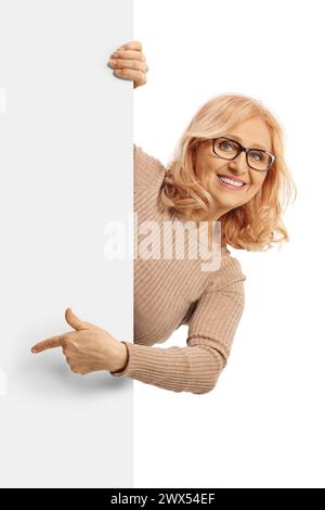 Donna di mezza età che sbircia da dietro un pannello bianco vuoto e punta isolata su sfondo bianco Foto Stock