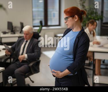 Una donna incinta soffre di dolore e tiene lo stomaco in piedi nel mezzo dell'ufficio accanto ai suoi colleghi. Foto Stock