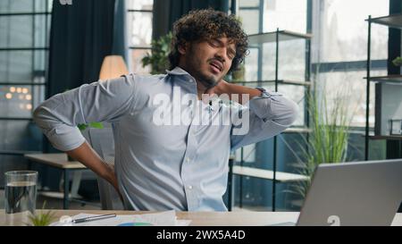 Malato uomo arabo-indiano latino-americano esausto sente dolore alla schiena dopo il lavoro del portatile male male ragazzo uomo di ufficio che massaggia doloroso collo Foto Stock