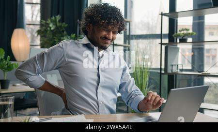 Malato uomo arabo-indiano latino-americano esausto sente dolore alla schiena dopo il lavoro del portatile male male ragazzo uomo di ufficio che massaggia doloroso collo Foto Stock