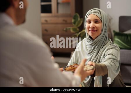 Spalla di una allegra donna musulmana che indossa l'hijab passando piatto con noci al suo parente maschio durante la cena di famiglia Foto Stock