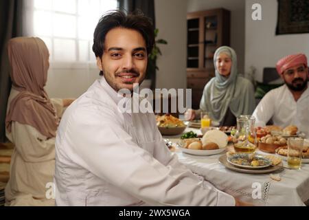 Ritratto di un giovane musulmano felice che indossa la kandora seduta al tavolo festivo con la sua famiglia su Eid al-Fitr che sorride alla macchina fotografica Foto Stock