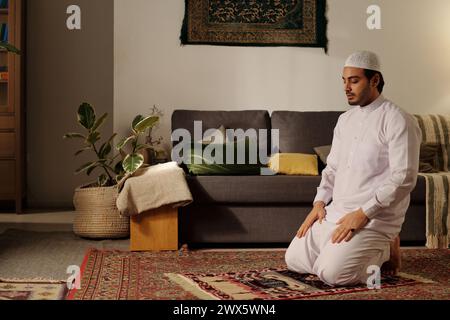 Giovane musulmano mediorientale seduto su un tappeto di preghiera con gli occhi chiusi facendo namaz nel soggiorno di casa Foto Stock