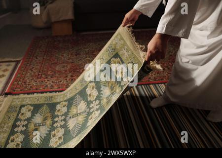 Uomo musulmano irriconoscibile che posiziona un tappeto di preghiera sul pavimento nel soggiorno di casa prima del namaz, copia spazio Foto Stock