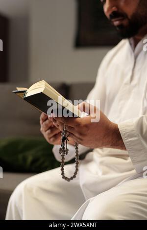 Ripresa selettiva verticale di un uomo mediorientale barbuto che tiene in mano un libro del Corano che legge la misbaha Foto Stock