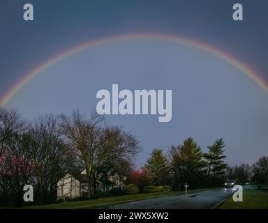 Strada suburbana del Midwest la sera delle piogge all'inizio della primavera con l'arcobaleno nel cielo; auto in lontananza Foto Stock