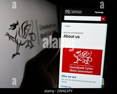 Persona che tiene un cellulare con pagina web del governo gallese (Llywodraeth Cymru) davanti al logo. Messa a fuoco al centro del display del telefono. Foto Stock
