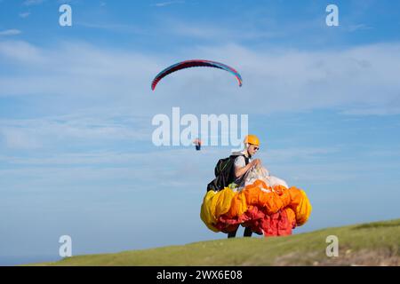 Uomo con il parapendio piegato nel cavolfiore e un altro parapendio volando dietro Foto Stock