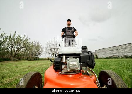 Il giardiniere professionista in abbigliamento protettivo taglia prati verdi utilizzando un moderno rasaerba a benzina senza fili nel cortile posteriore. Giardino stagionale desi Foto Stock