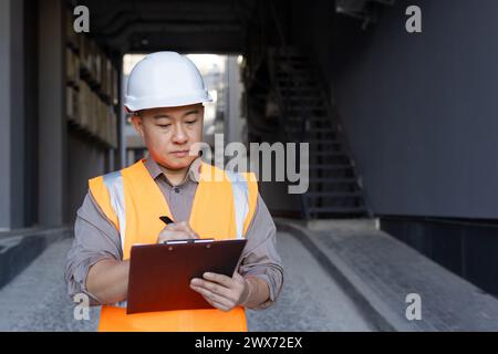 Concentrato e serio giovane architetto asiatico maschio, costruttore. l'ingegnere si trova all'esterno con un elmetto e un giubbotto e prende appunti nei documenti, conduce ispezioni, redige un piano di progetto. Foto Stock