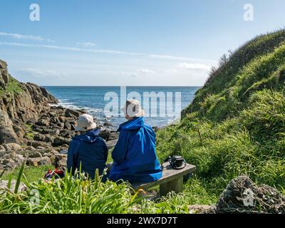 Due camminatori lungo il percorso costiero seduti sul sedile che si affaccia sulla baia di Porthgwarra sul sentiero costiero della Cornovaglia meridionale a marzo, Cornovaglia, Inghilterra, Regno Unito Foto Stock