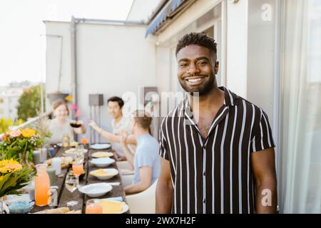 Ritratto di un giovane sorridente che indossa una camicia a righe con gli amici mentre cena in balcone Foto Stock