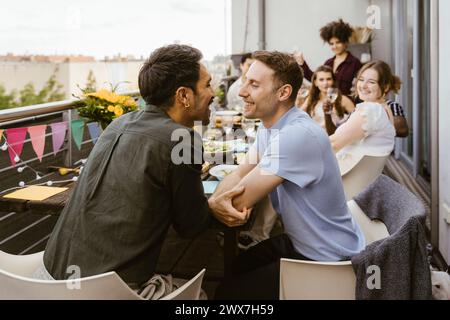 Romantica coppia gay che si guardano mentre si siede con gli amici a cena sul balcone Foto Stock