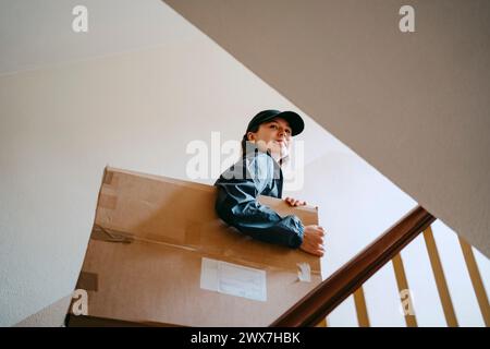 Vista ad angolo basso della persona che sorride al parto che tiene in mano una scatola di cartone mentre si sale sulle scale dell'edificio Foto Stock