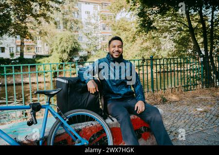 Ritratto di un uomo felice che consegna il cibo seduto sulla panchina in bicicletta al parco Foto Stock