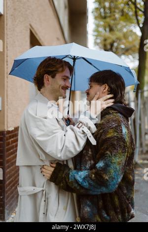 Vista laterale di una giovane coppia gay romantica in piedi sotto l'ombrello sulla strada Foto Stock