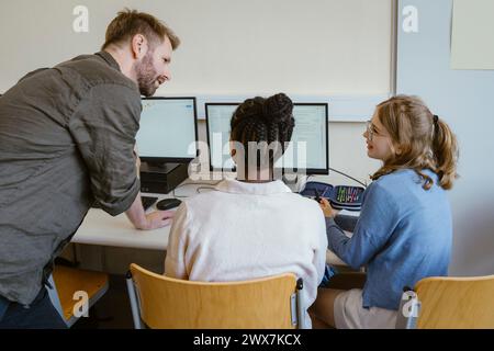 Insegnante di sesso maschile che discute con studentesse sedute su sedie in classe di computer a scuola Foto Stock