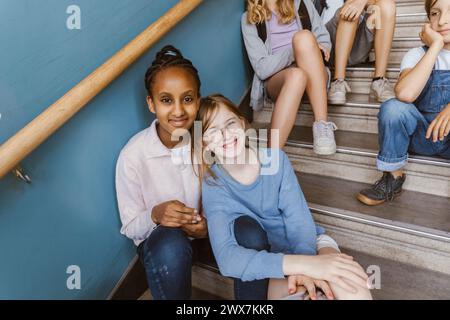 Ritratto di amiche multirazziali sorridenti sedute su una scala nell'edificio scolastico Foto Stock