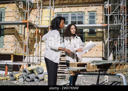 Donna professionista del design che mostra i progetti al cliente mentre si trova di fronte all'appartamento in costruzione Foto Stock