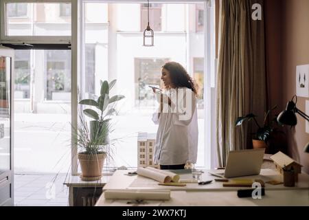 Design femminile sorridente professionale che tiene in mano la tazza mentre parla tramite il telefono con vivavoce vicino alla scrivania in ufficio Foto Stock