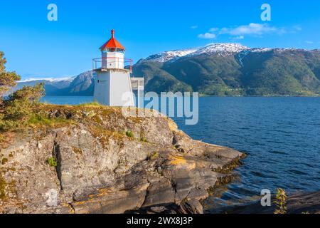 Faro su una collina rocciosa lungo la costa della pittoresca cittadina norvegese Leikanger lungo il Sognefjord norvegese con cieli blu e verdi montagne innevate Foto Stock