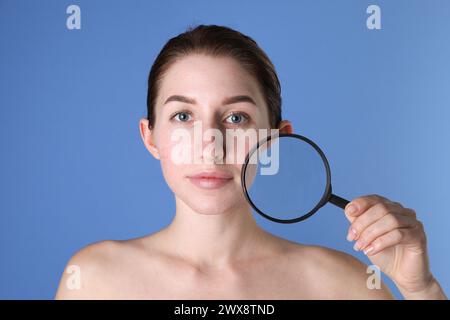 Giovane donna con problemi di acne tenendo la lente di ingrandimento vicino alla pelle su sfondo blu Foto Stock