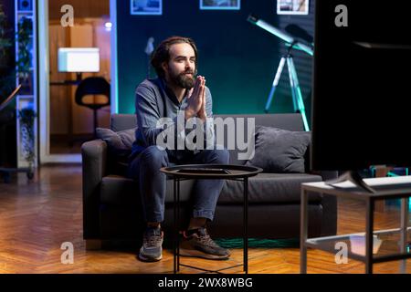 Uomo fiducioso che prega perché la squadra preferita vinca mentre guarda gli eventi sportivi in diretta trasmessi in TV. Tifoso seduto sul divano di casa implorando i gesti della mano, godendo di una competizione calcistica Foto Stock