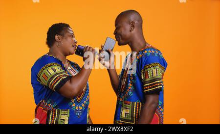 Felice coppia afroamericana che canta canzoni e registra la propria voce sul microfono dello smartphone, godendosi una divertente sessione di karaoke. Allegra gente etnica in abiti tradizionali registra melodie. Foto Stock