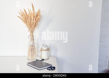 Bellissimo bouquet di fiori d'erba di pampas essiccati, fiori di canna in un vaso di vetro contro la parete bianca, candela, taccuino. Sfondo con spazio di copia, vuoto. Modello, mock-up per il tuo design, poster, arte. Foto Stock