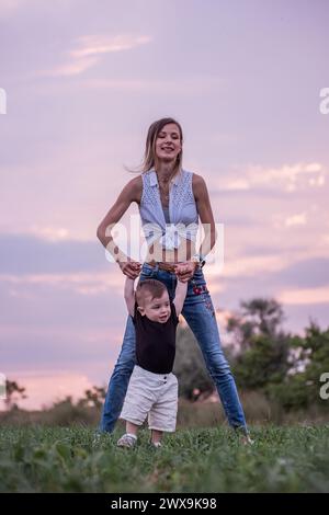 La donna solleva il bambino in alto in aria sullo sfondo del cielo viola del tramonto, un momento di pura gioia. Madre e figlio piccolo che giocano con me verde Foto Stock