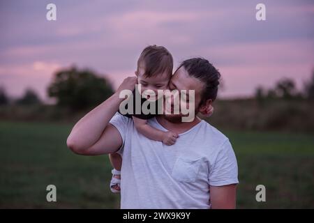 Scena emozionante di padre della diversità che porta un figlio sorridente sulle spalle al crepuscolo, mostrando affetto e legami familiari. Tenero padre figlio abbraccio. Foto Stock