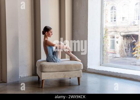 La giovane ballerina in abito blu si siede accanto alla finestra in posa serena, riflettendo tranquillamente prima della lezione di balletto o dello spettacolo. Young Dancers Contemplative Mo Foto Stock