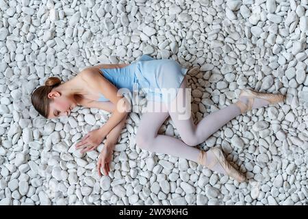 Ballerina eterea con vista dall'alto e cordoncino blu, collant appoggiati con grazia sullo sfondo di lisci ciottoli bianchi, giustapposizione di delicata forma umana ag Foto Stock