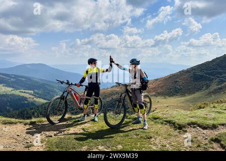 Due ciclisti uomini che guidano biciclette elettriche all'aperto. Turisti maschi che riposano sulla cima della collina, si danno cinque alti l'uno all'altro, si godono il bellissimo paesaggio montano, indossando casco e zaino. Foto Stock