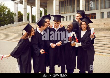 Gruppo di laureati multirazziali felici e risi che abbracciano all'aperto con un diploma. Foto Stock