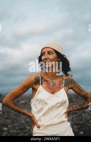 Giovane donna felice tatuata in un vestito su una spiaggia rocciosa ventosa. Foto Stock