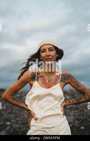 Giovane donna felice tatuata in un vestito su una spiaggia rocciosa ventosa. Foto Stock