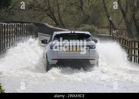 Lacock, Wiltshire, Regno Unito. 29 marzo 2024. Il fiume Avon a Lacock ha fatto scoppiare le sue sponde e ha inondato la strada dentro e fuori il villaggio. Il Consiglio è di non guidare attraverso l'acqua piena, ma la strada è percorribile se si vuole fare un tuffo. Crediti: JMF News/Alamy Live News Foto Stock