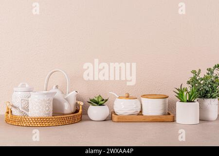 Set da tè in ceramica bianca su vassoio rotondo in vimini tra le piante da interno sul ripiano della cucina. Sfondo della cucina in colori chiari. Uno spazio di copia. Vista frontale Foto Stock