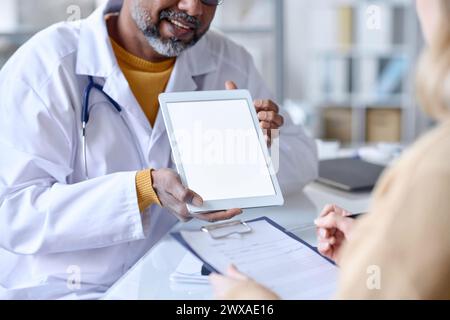 Primo piano di un medico sorridente che mostra una compressa con schermo bianco vuoto al paziente durante la consultazione nello spazio di copia della clinica Foto Stock